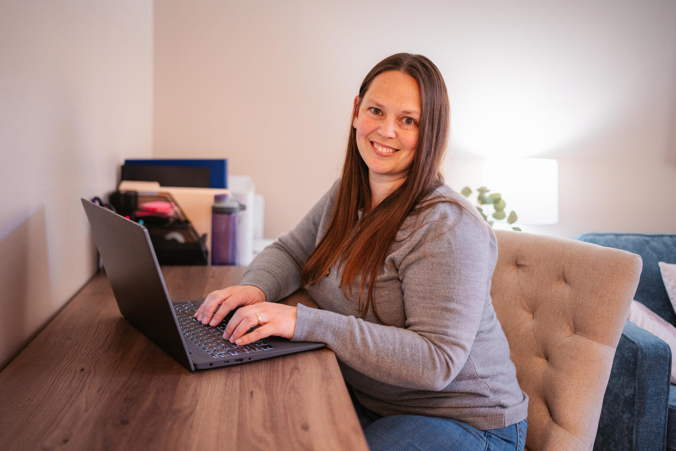 Smiling woman with long brown hair typing on a laptop at a wooden desk in a cozy home office.